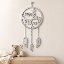 A silver dreamcatcher wall hanging with the words 'Dream big little one' and three hanging feather shapes, displayed on a beige wall above a wooden surface with a crocheted teddy bear and a small vase.